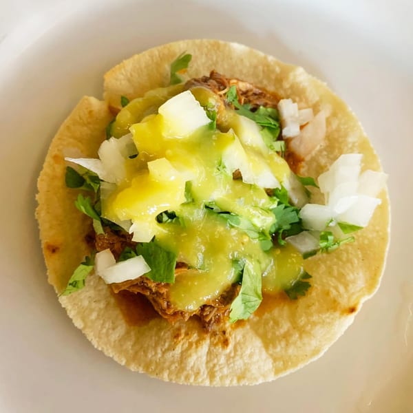 A single street taco with meat, green salsa, diced onions, and cilantro on a white plate.