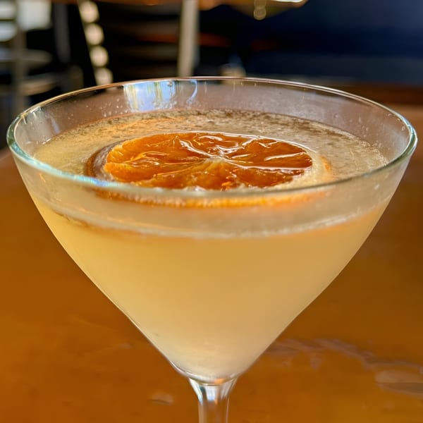 Close-up of a martini glass with a light yellow cocktail garnished with a dried orange slice.