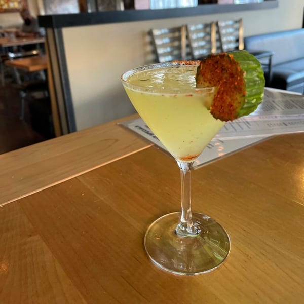 A green cocktail in a martini glass, garnished with a cucumber slice and chili powder.