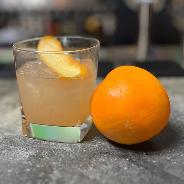 A cocktail with an orange peel garnish sits next to a whole orange on a dark surface.
