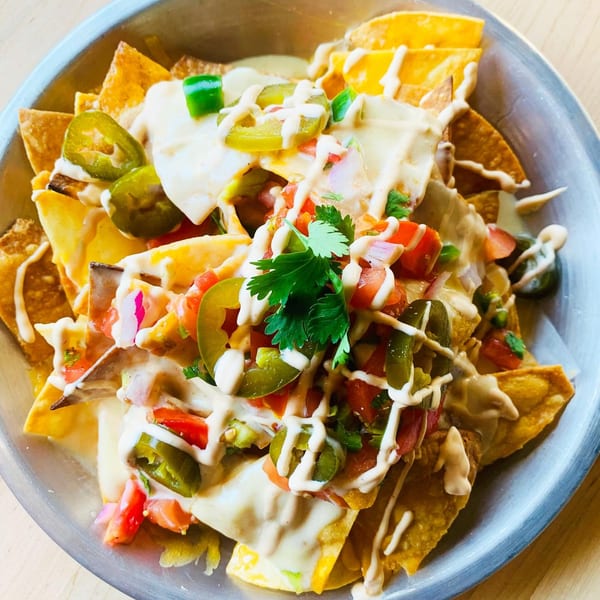 A close-up of a metal bowl filled with nachos, topped with cheese sauce, jalapeños, pico de gallo, and cilantro.