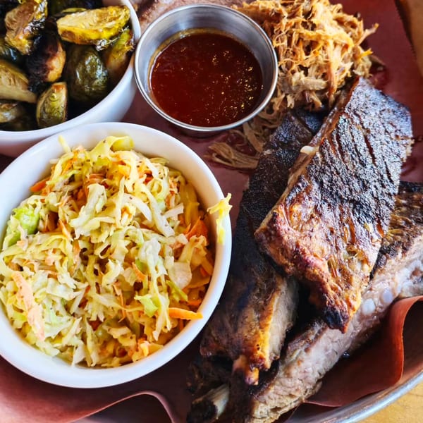 A platter of grilled ribs, pulled pork, coleslaw, roasted Brussels sprouts, and BBQ sauce.