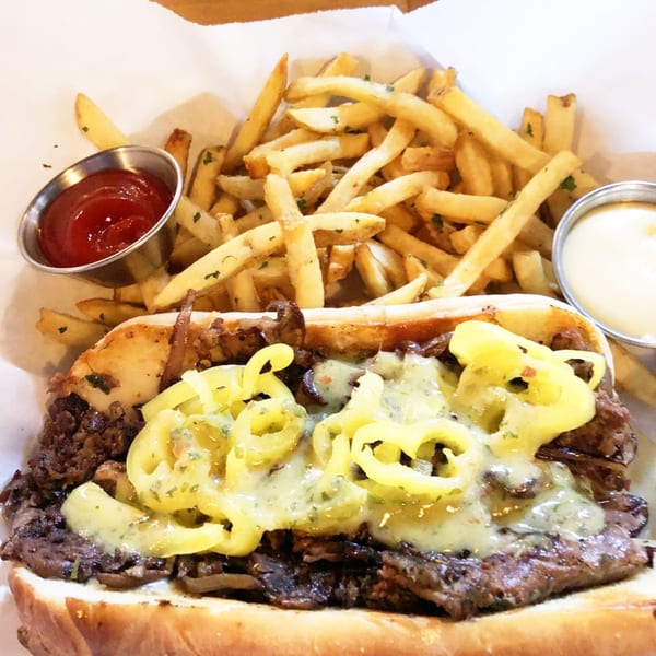 A cheesesteak sandwich with peppers and fries, ketchup, and aioli on a white paper-lined tray.
