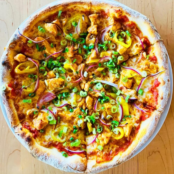 A whole BBQ chicken pizza with red onions, banana peppers, and green onions on a wooden table.