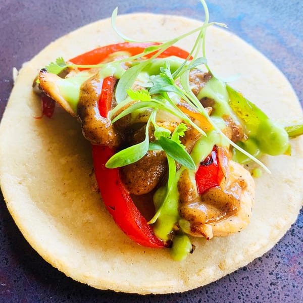 A chicken fajita taco with red and green peppers, drizzled with two sauces and garnished with microgreens.